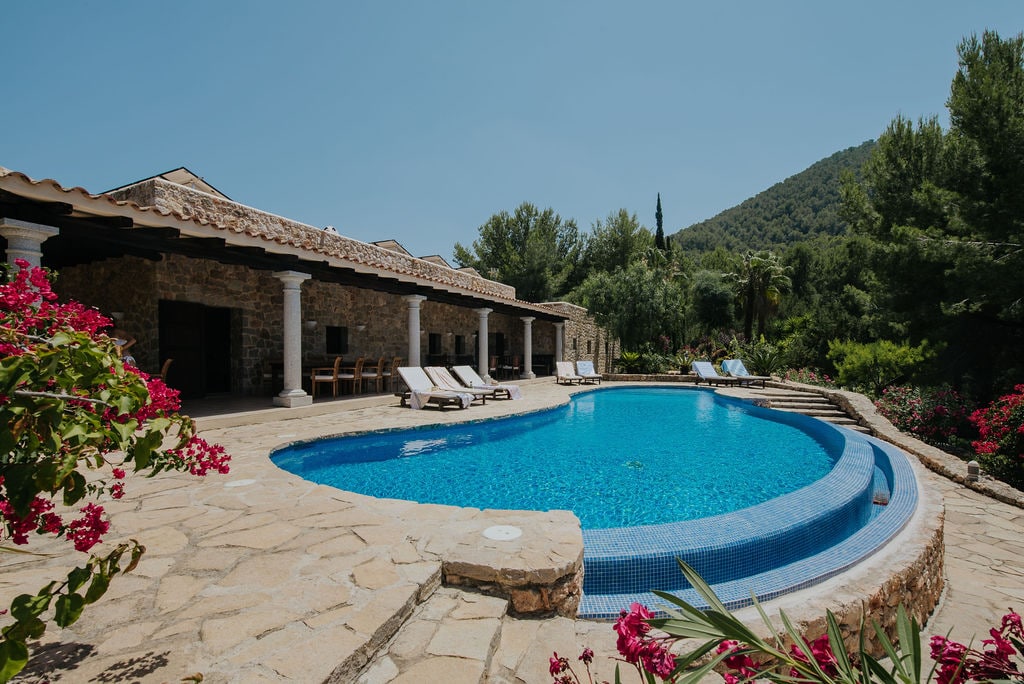A stone terrace surrounds a large, inviting pool, framed by vibrant flowers and lush greenery. Sun loungers are positioned around the pool, offering a serene spot for relaxation. In the background, a hillside of trees adds to the tranquil atmosphere.