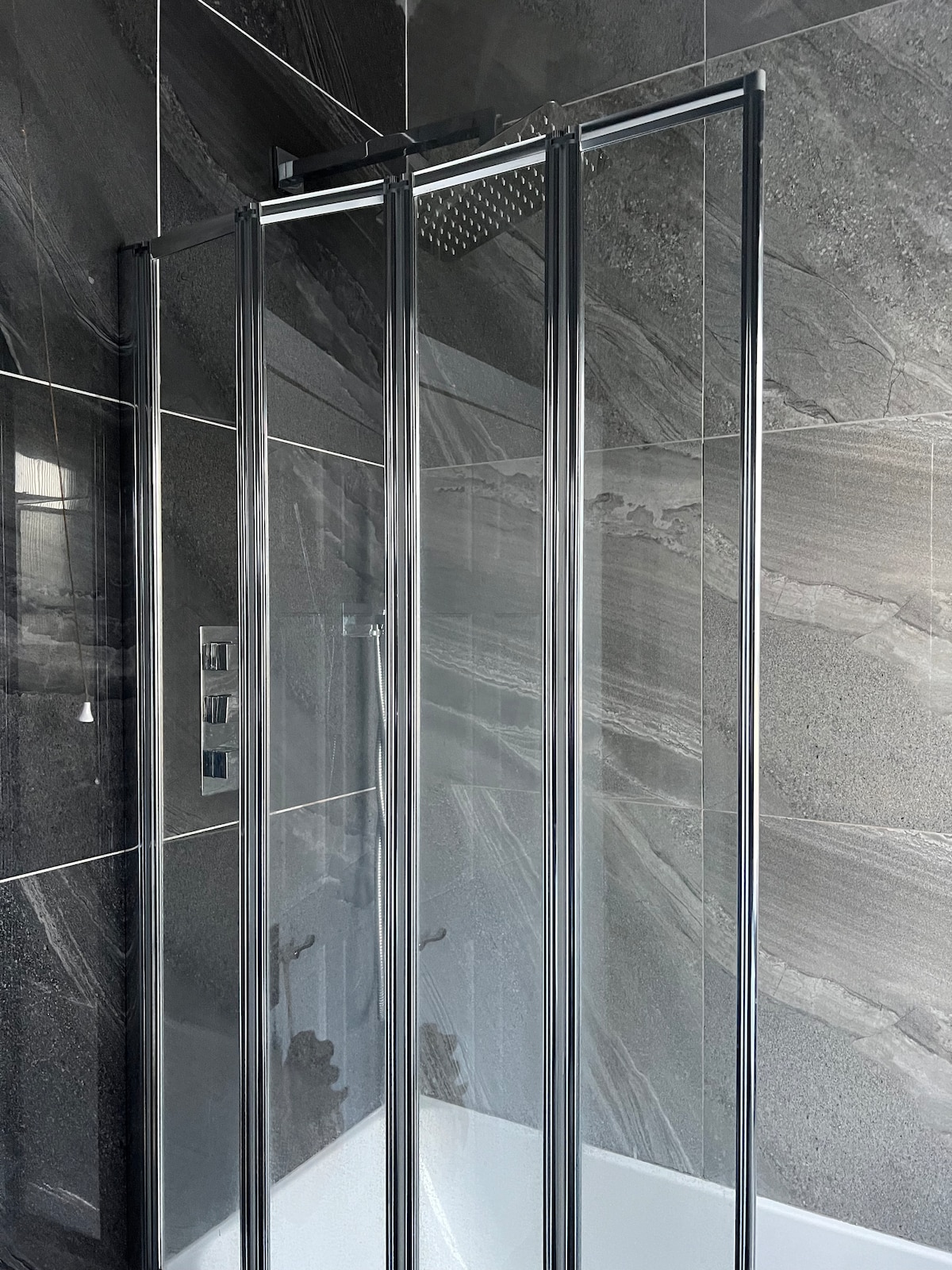 A modern shower enclosure features sliding glass doors, allowing for easy access. The walls are adorned with sleek dark tiles that create a contemporary aesthetic. A rainfall showerhead is mounted above, enhancing the overall minimalist design of the space.