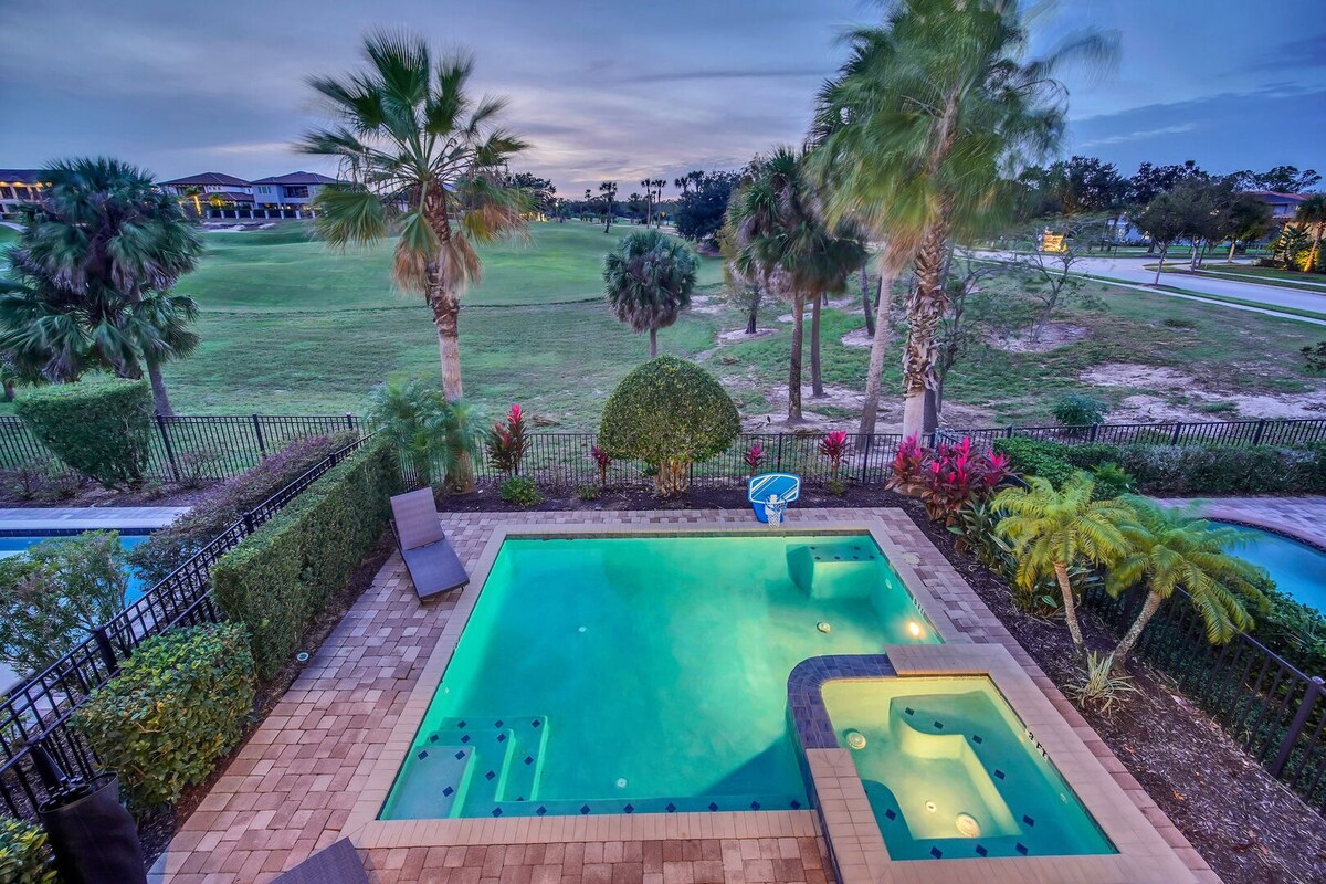 A private pool and spa are surrounded by tropical landscaping, featuring palm trees and colorful shrubs. The pool area is paved with decorative tiles, and the water's surface reflects the evening sky. Nearby golf course views are visible beyond the pool's enclosure.