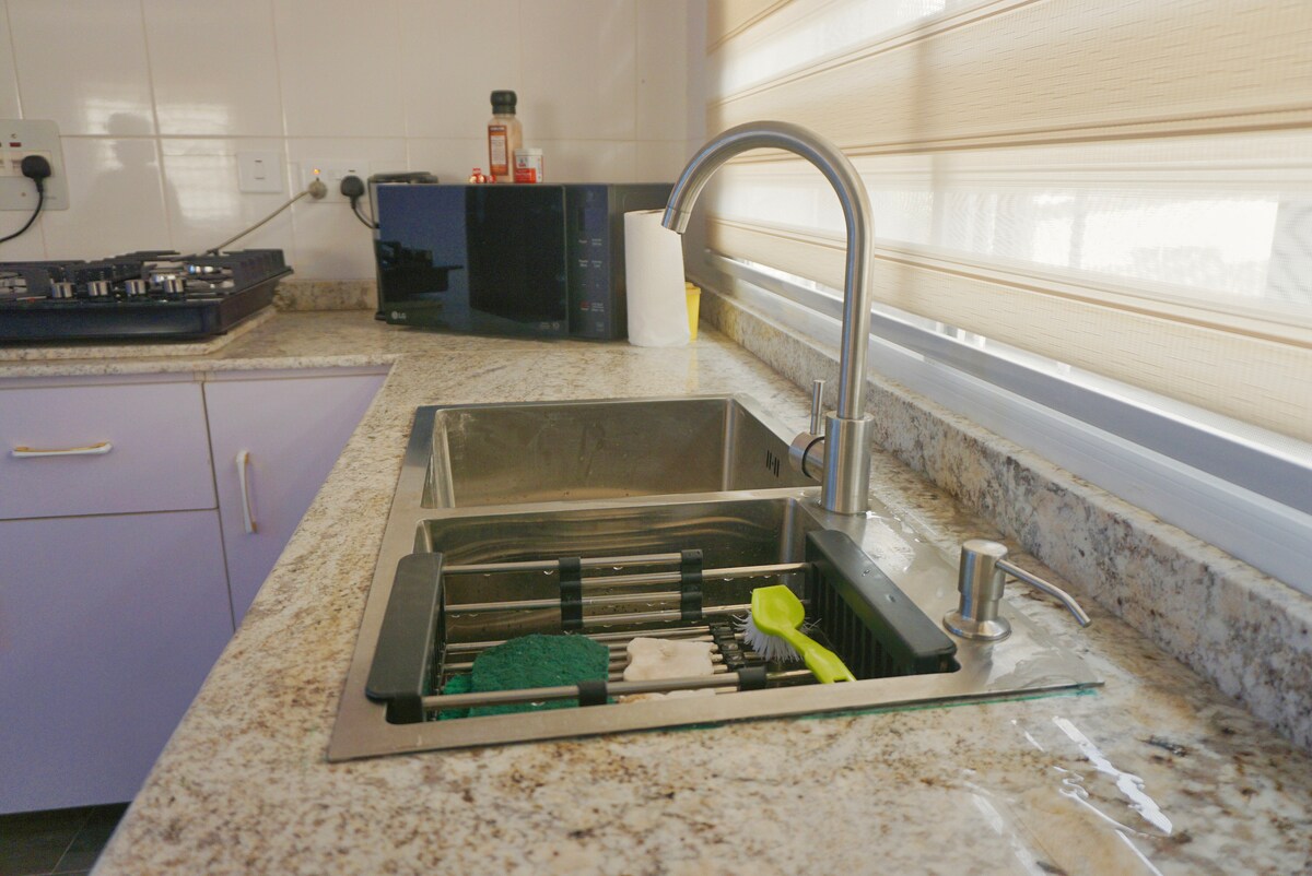 A modern kitchen sink area is presented with a stainless steel double basin. A sleek faucet stands prominently above the sink, while a dish drying rack is placed neatly beside. The countertops feature a light granite surface, complemented by a blending of kitchen appliances.