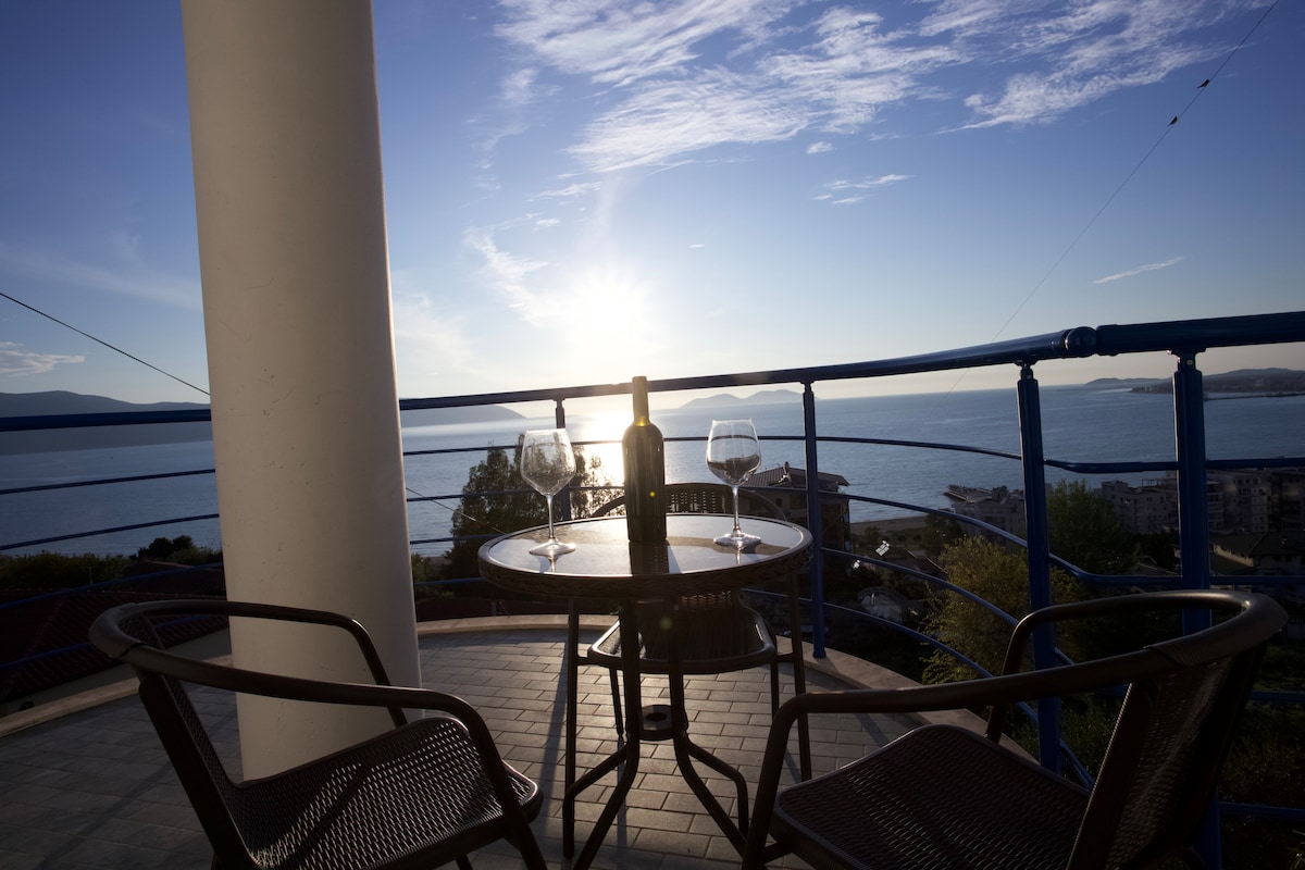 A balcony table is set for two, featuring two glasses and a wine bottle. The view includes a tranquil sea under a clear sky, with the sun rising on the horizon, casting soft light over the scene.