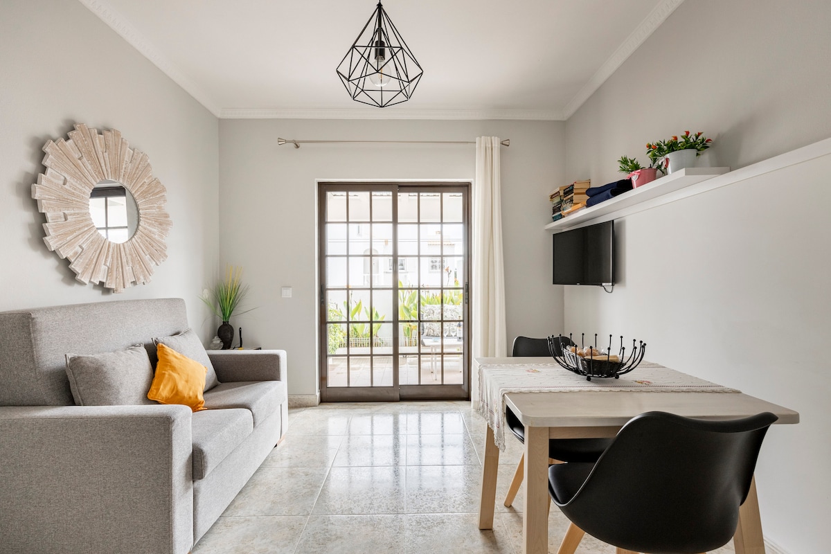 A light-filled living space features a comfortable grey sofa adorned with an orange cushion. A minimalist coffee table is placed in front, accompanied by black chairs. Large glass doors provide access to outdoor views, while a decorative mirror enhances the room's aesthetic.