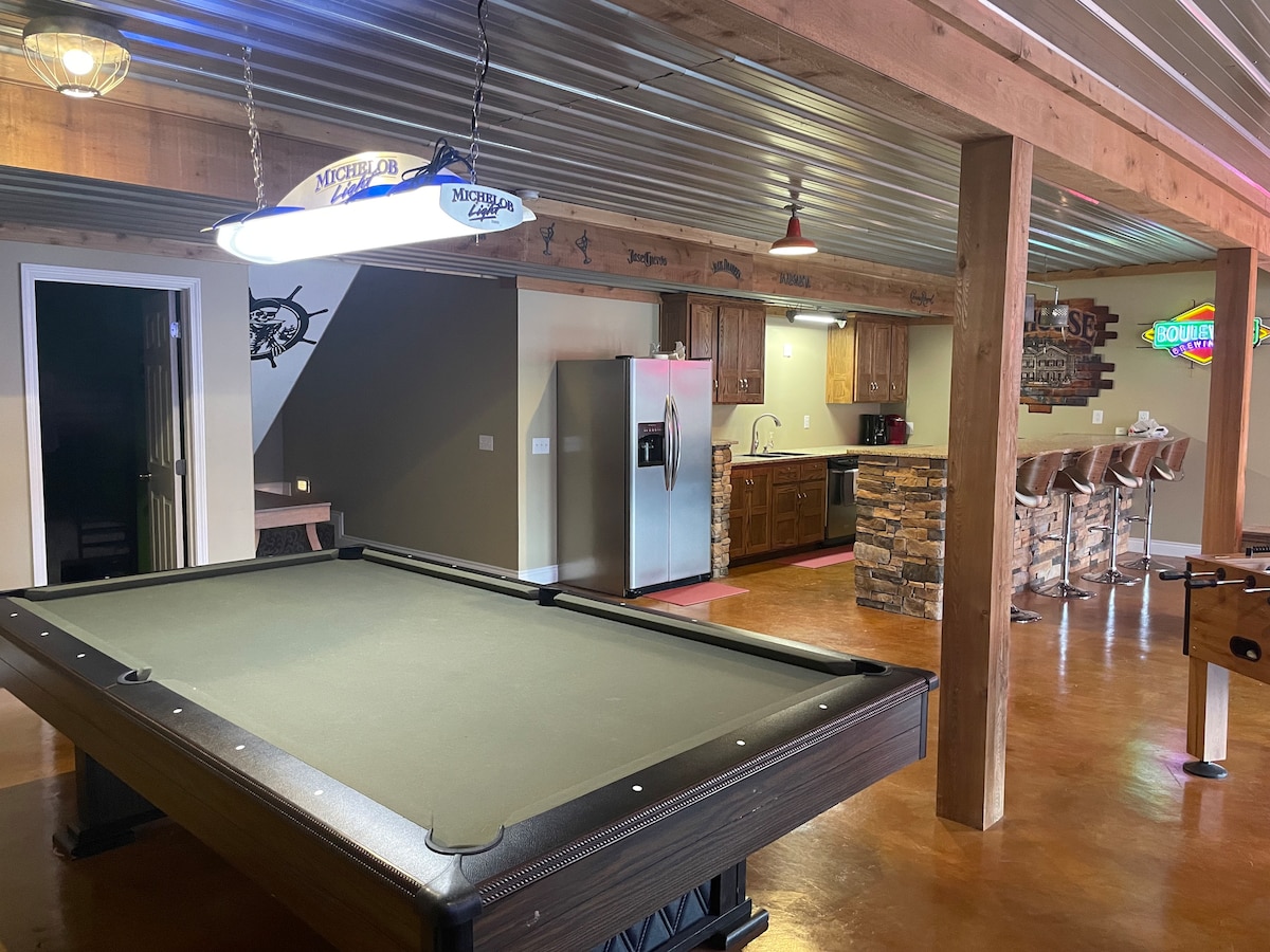 A spacious game room features a billiards table at its center, surrounded by wooden support beams. Adjacent to it, a fully equipped kitchen is visible, with modern appliances and a bar area consisting of several high chairs. Soft lighting highlights the inviting atmosphere.