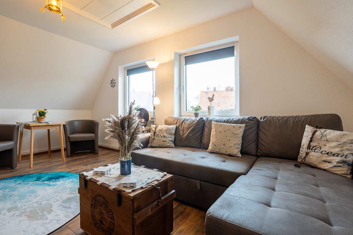 The living area features a comfortable gray sectional sofa adorned with decorative pillows. A wooden coffee table sits on a blue patterned rug, and two chairs are positioned around a small dining table near the window. Natural light fills the space, accompanied by a floor lamp.