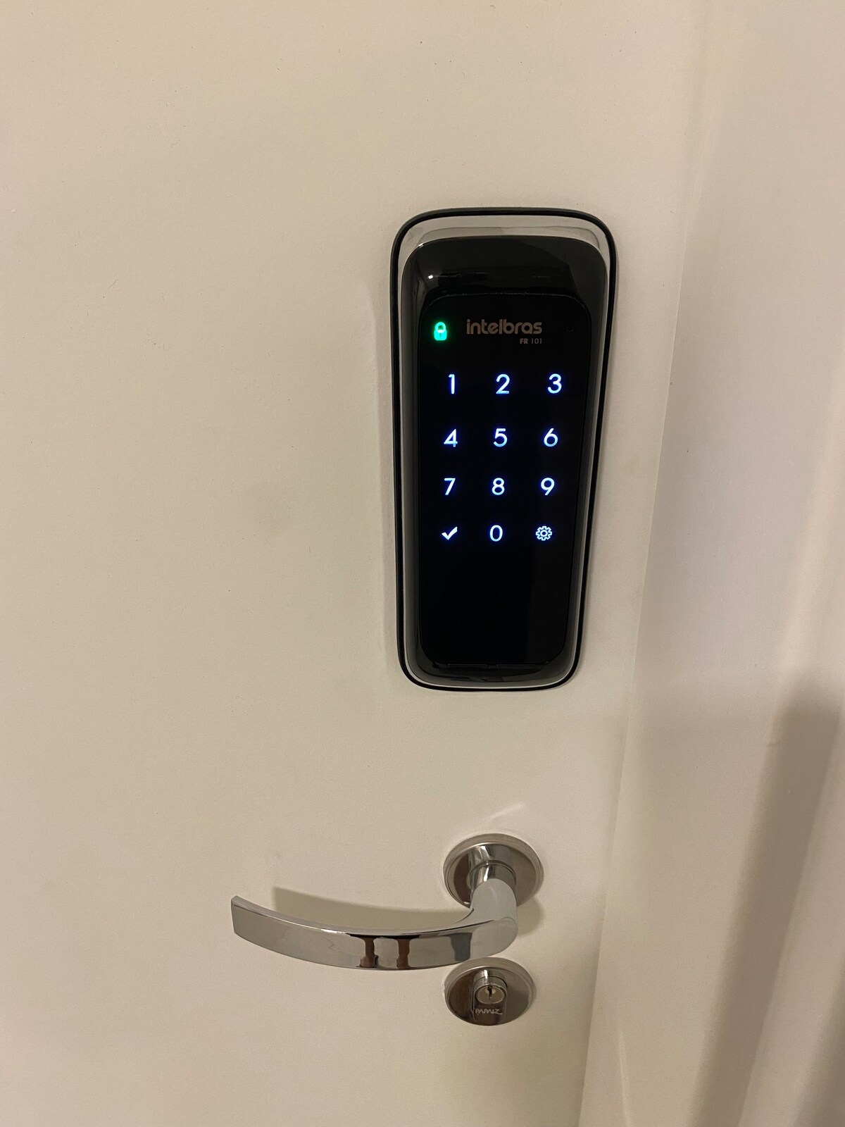 A modern electronic keypad is mounted on a smooth white door, featuring illuminated numbers and a prominent indicator light. A sleek, metallic door handle is positioned beneath the keypad, emphasizing the contemporary design of the entrance.