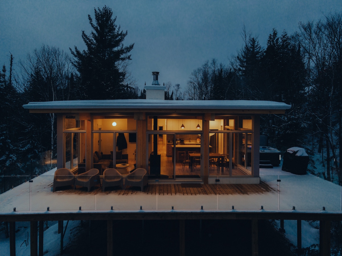 The chalet features a modern design surrounded by snow, with large glass doors offering views into the warm, illuminated interior. A spacious deck holds comfortable outdoor seating, and a spa area is visible in the background, all set against a tranquil wintry landscape.