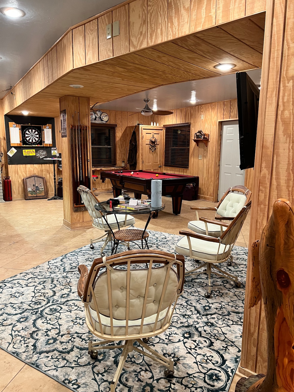 A cozy recreation area features a pool table and a dartboard, surrounded by warm wooden paneling. Comfortable seating includes metal-framed chairs on a decorative rug, creating an inviting space for relaxation and entertainment. Soft lighting enhances the room's welcoming atmosphere.
