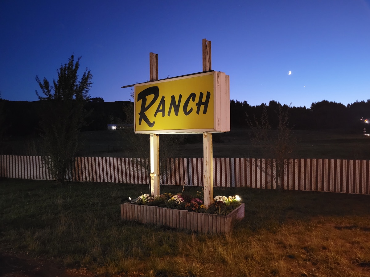 A large, vintage sign displaying the word 'Ranch' in bold yellow letters is set against a twilight sky. The sign is flanked by simple landscaping with low flowers illuminated by ground-level lights, providing a welcoming entrance to the property.