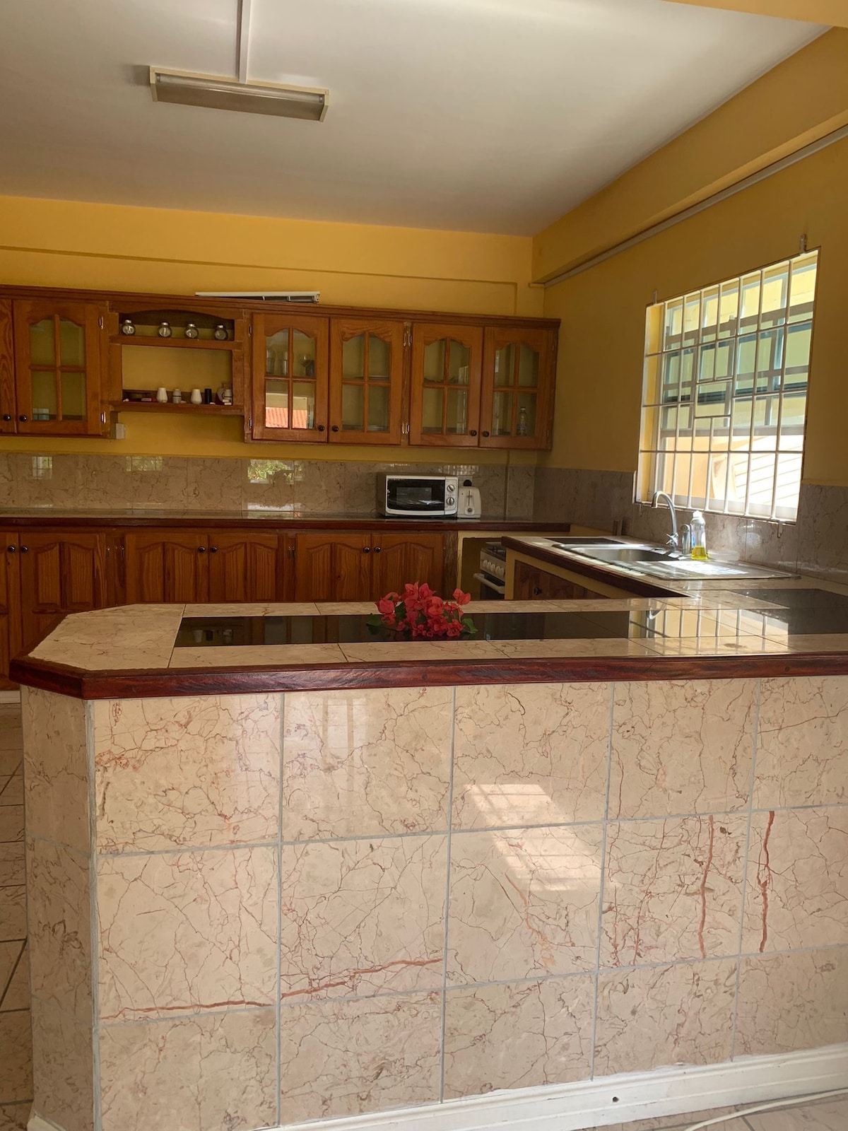 A spacious kitchen features wooden cabinetry and marble countertops, providing ample storage and workspace. Natural light enters through windows, highlighting the nearby sink and microwave. A small arrangement of flowers is displayed on the counter, adding a touch of color to the neutral tones.