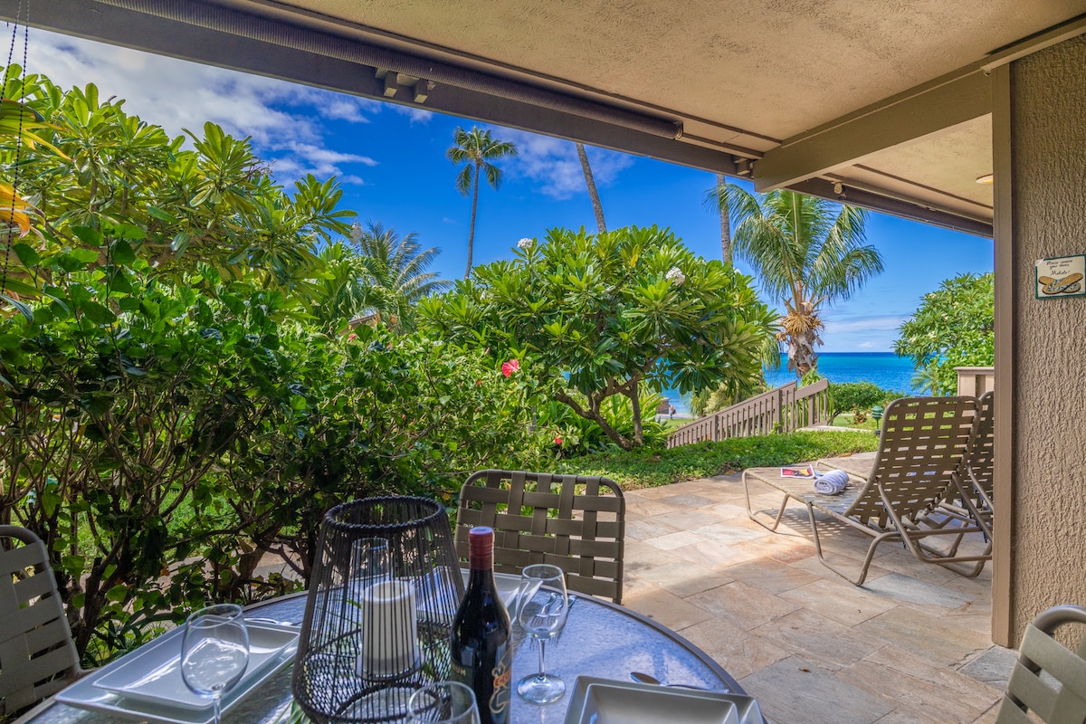 A covered lanai is shown, featuring a dining table set for four with glasses and wine. Lush greenery surrounds the space, with palm trees visible in the background. The tranquil ocean can be seen in the distance under a clear blue sky.