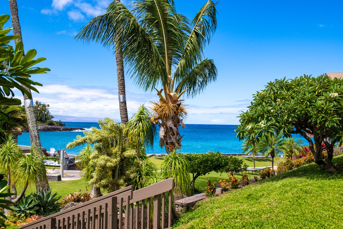 Lush green lawns surround the manicured gardens, while palm trees sway gently in the ocean breeze. A vibrant blue ocean is visible in the background under a clear sky, inviting relaxation and leisure by the water.