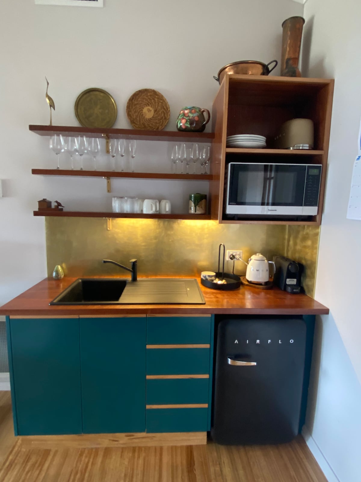 A compact kitchenette features a sleek gold backsplash and wooden countertop, highlighting a modern sink and an array of dishes. Open shelving holds glassware, while a microwave and coffee making facilities are integrated into the cabinetry. A small refrigerator is positioned beneath the counter.