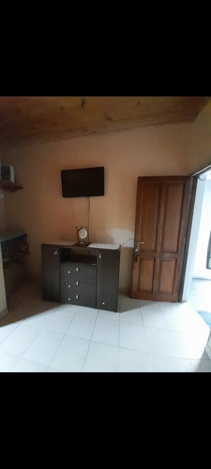A compact room features a wooden cabinet with multiple drawers and a flat-screen television mounted on the wall above. A door leading outside is visible, alongside smooth tiled flooring. Natural light enters the space, creating a bright and functional atmosphere.