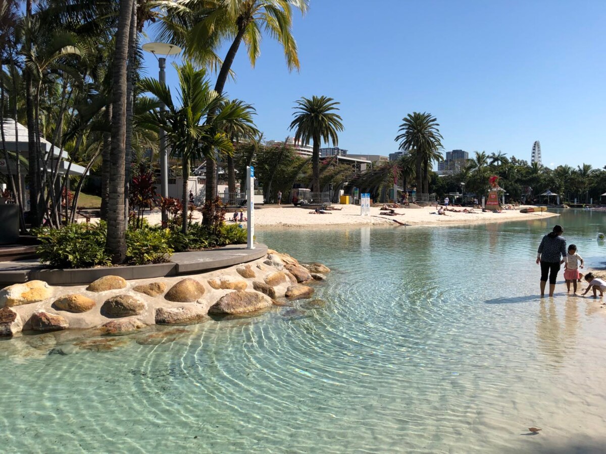 A serene beach area is bordered by palm trees and gentle waves, with sandy shores visible in the background. Adults and children can be seen enjoying the water, while inviting lounge chairs are arranged nearby for relaxation.