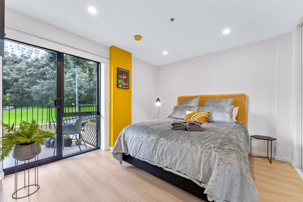 A well-lit bedroom features a spacious bed with textured grey bedding and coordinating cushions. A small side table stands next to the bed, while large glass doors open to a balcony overlooking greenery. A vibrant yellow wall adds a fresh accent to the room.