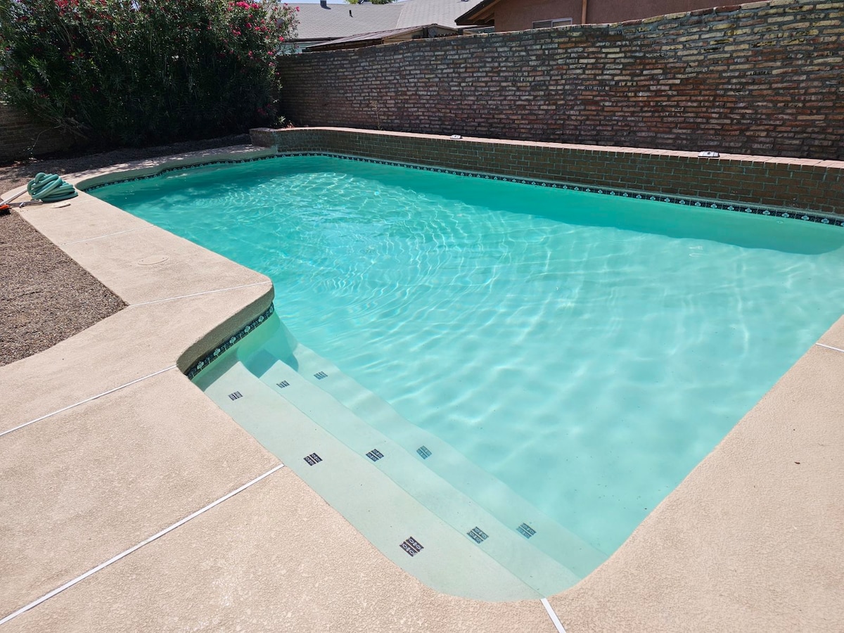 A heated swimming pool is surrounded by concrete decking, featuring a gentle slope for easy entry. The clear turquoise water reflects sunlight, creating a serene setting. Lush greenery can be seen along one side, with a low brick wall providing privacy.
