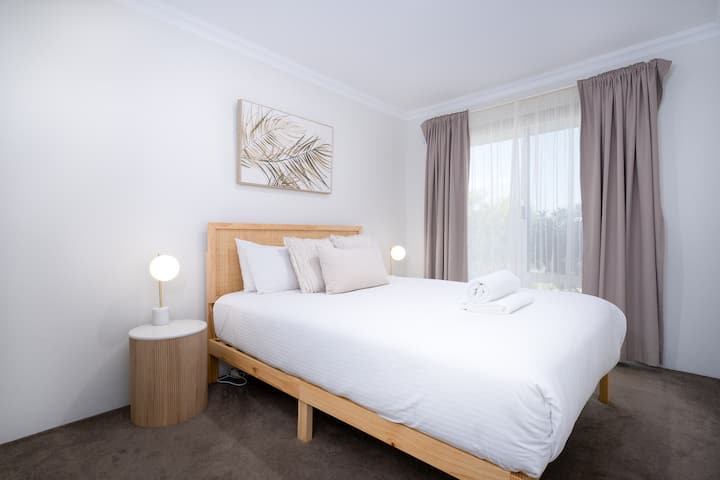 Chambre lumineuse et minimaliste avec un lit queen size confortable avec des draps blancs frais, deux tables de chevet avec des lampes modernes et des rideaux beige doux. Décoré avec de l'art mural apaisant pour une atmosphère sereine.
