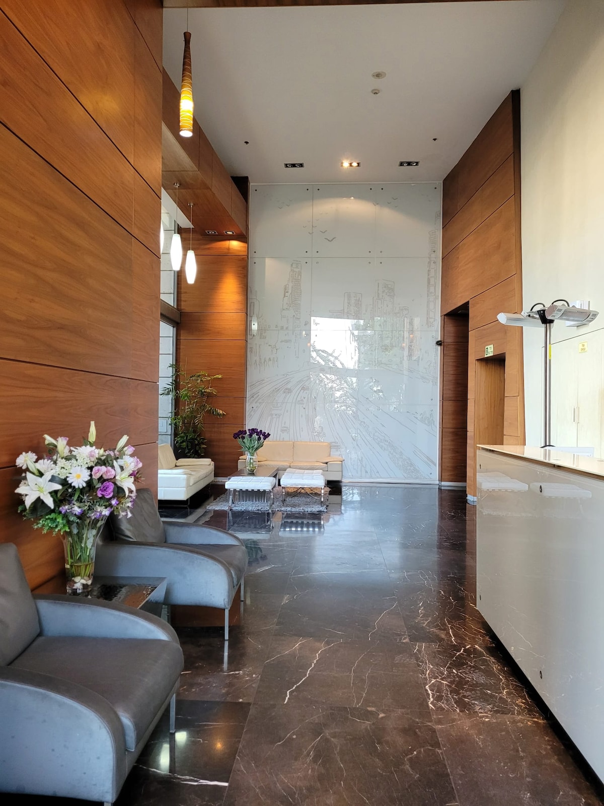 A sleek entrance area features warm wood paneling and polished marble floors. Comfortable seating is arranged beside a modern reception desk. Elegant flower arrangements add a touch of color. Natural light filters through large windows, creating an inviting atmosphere.