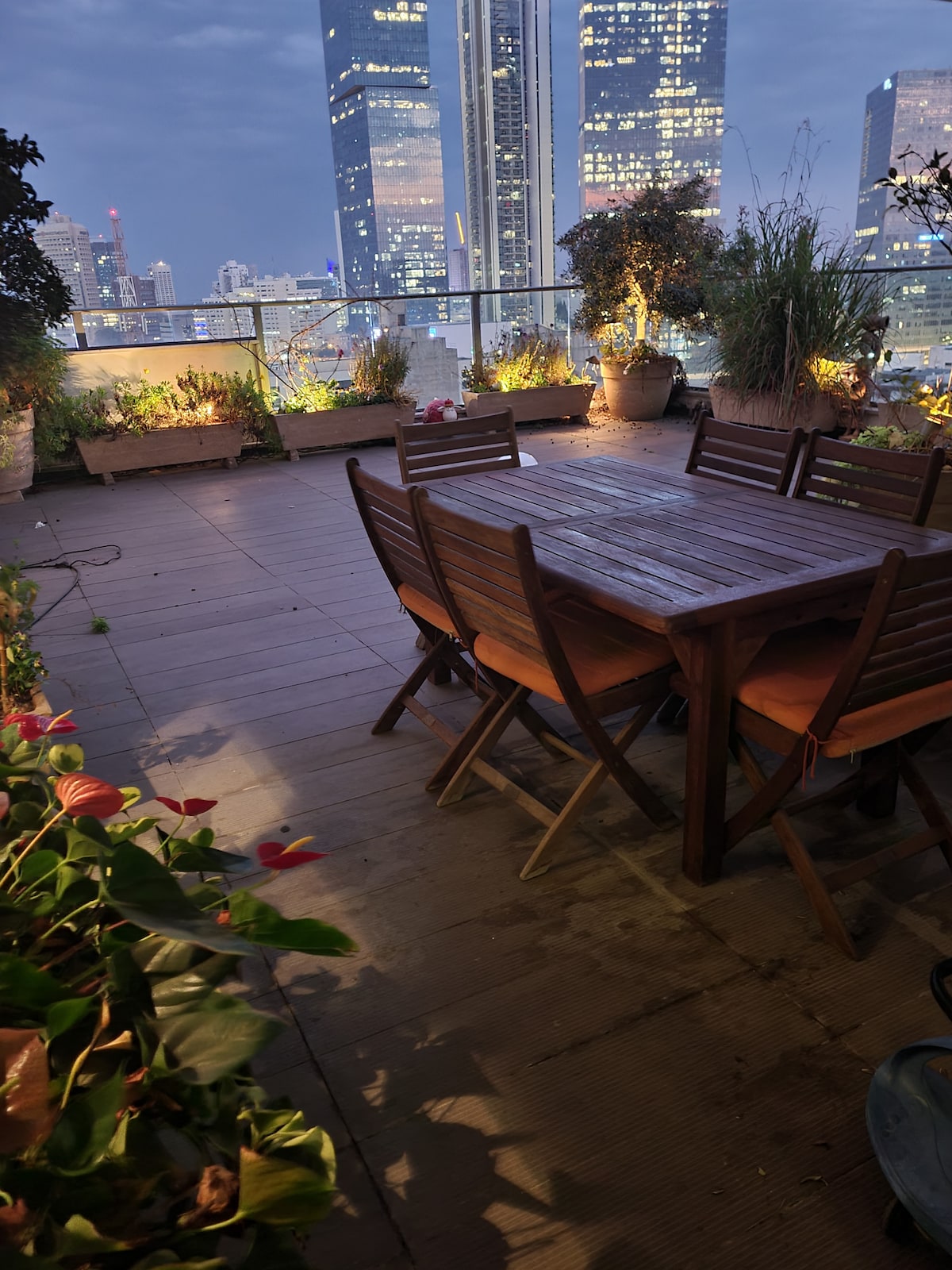 An outdoor terrace is presented, featuring a large wooden dining table surrounded by several chairs. Lush plants and flowers are arranged in pots along the edges, with city lights visible in the background, contributing to an urban atmosphere.