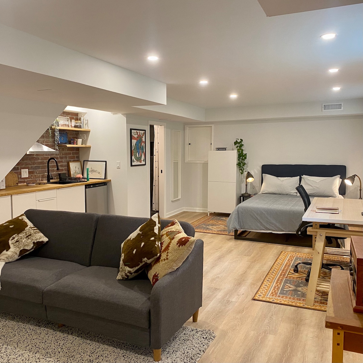 A well-lit studio apartment is shown with a combination of modern furniture. A gray sofa with accent pillows faces a bed dressed in neutral tones, adjacent to a dedicated workspace featuring a desk and ergonomic chair. Light wood flooring enhances the open layout.