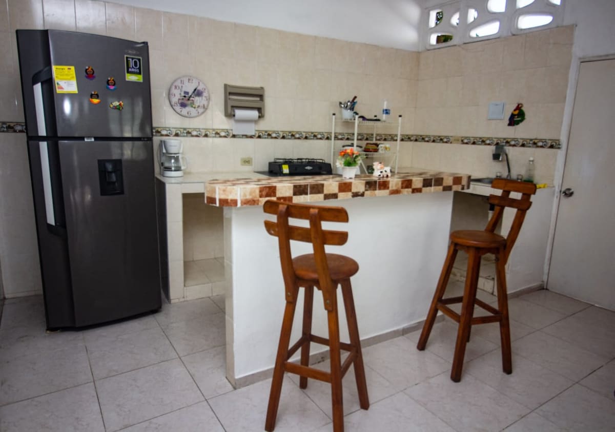 A functional kitchen space features light-colored tiled walls and a small bar area with two wooden stools. Included is a stainless steel refrigerator and a gas stove, with a coffee maker visible on the countertop. Decor elements such as a wall clock and a small vase add a welcoming touch.