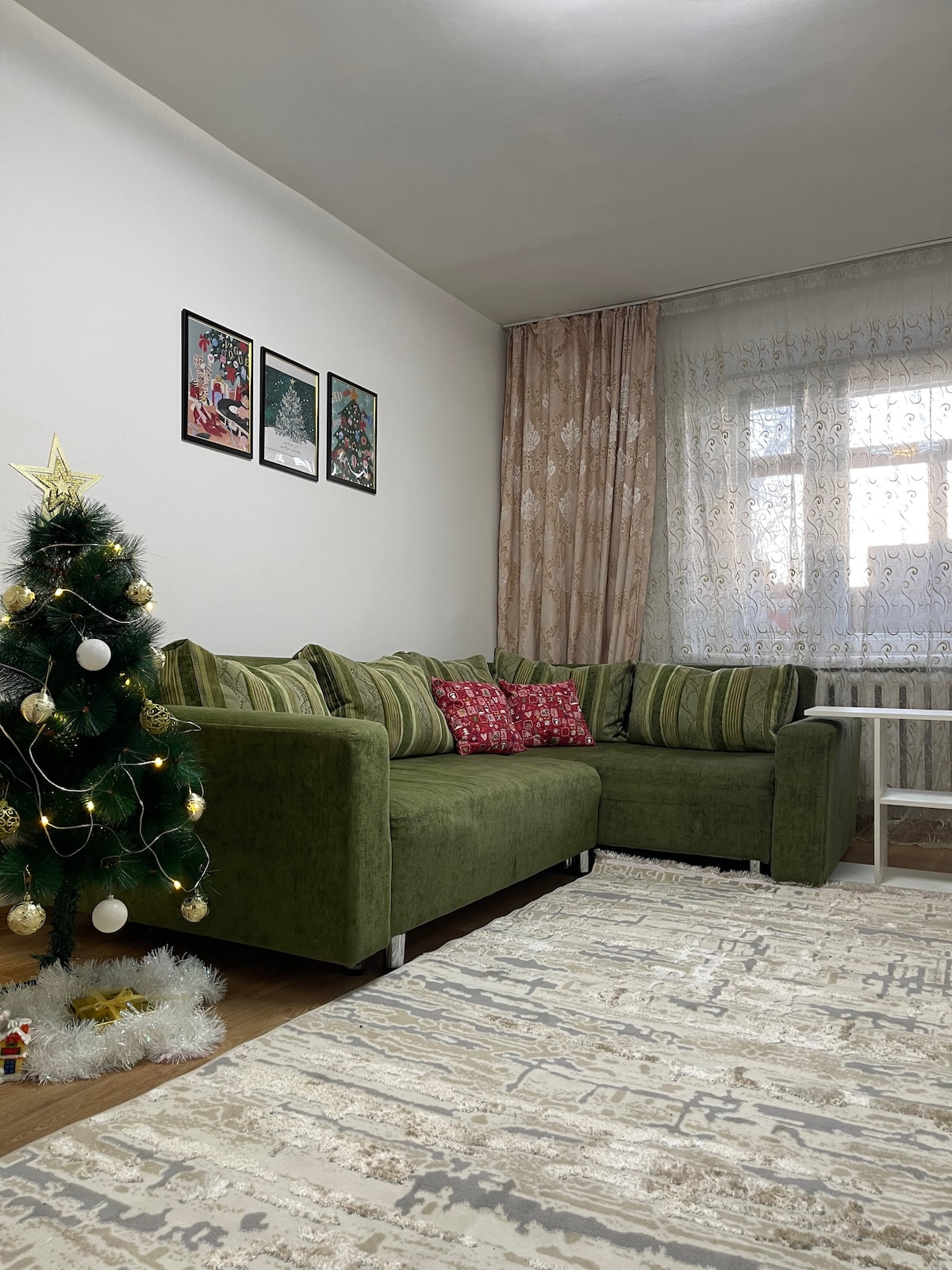 A cozy living area features a green sectional sofa adorned with colorful cushions. A small white table is positioned nearby. A festive Christmas tree with decorative lights and ornaments stands in the corner, adding a seasonal touch, while sheer curtains filter natural light from the window.
