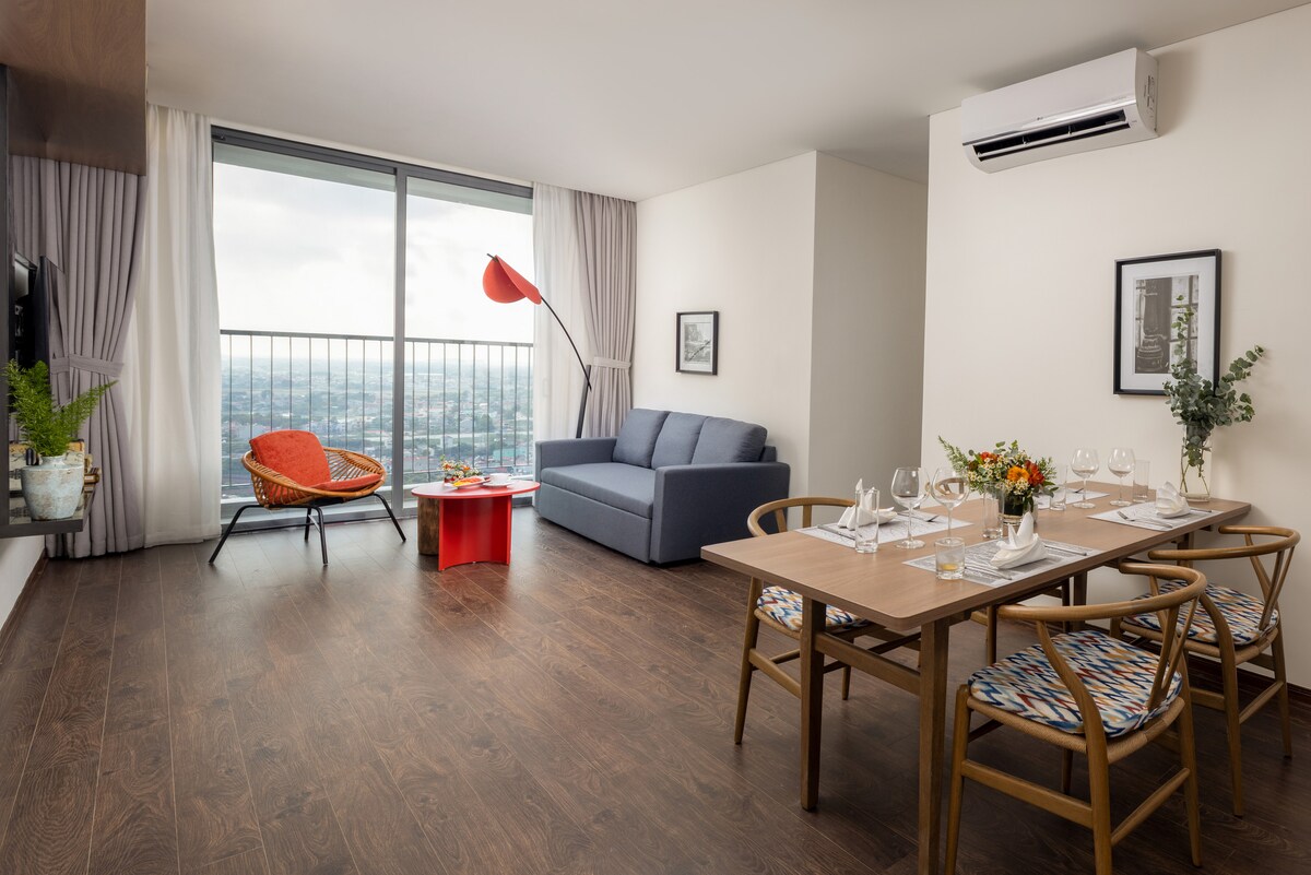 A modern living area showcases a blue sofa and an orange accent chair. A wooden dining table is set for four, with glassware and tableware arranged neatly. Floor-to-ceiling windows offer views of the surroundings, complemented by indoor plants and contemporary decor.