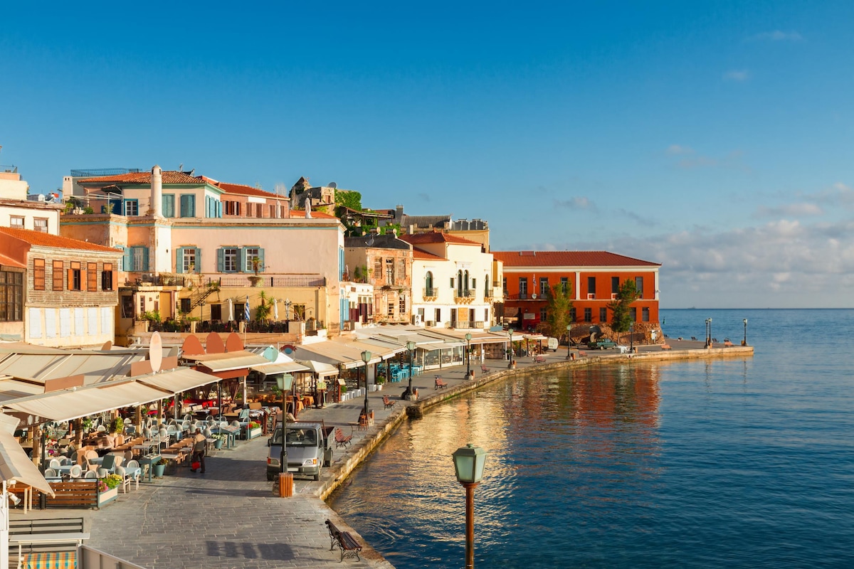 A waterfront scene features a promenade lined with cafes and restaurants, reflecting a local atmosphere. Colorful buildings with varying architectural styles rise above the sandy shore, while the calm sea extends to meet a clear blue sky, evoking a sense of tranquility.