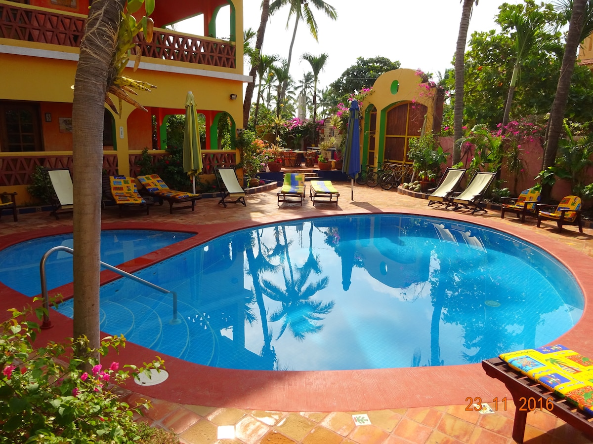 A curved swimming pool is surrounded by lush greenery and vibrant flowers. Loungers are arranged around the pool, inviting relaxation. Palm trees gently sway in the background, contributing to a serene atmosphere. Colorful patio chairs can be seen, adding an inviting touch to the outdoor space.