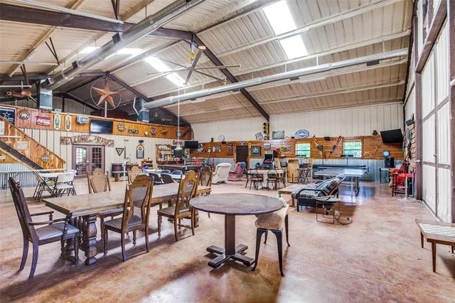 An expansive entertainment hall is showcased, featuring several seating arrangements including tables and chairs. High ceilings are adorned with exposed beams, while rustic decor enhances the space. A bar area and game stations are visible in the background, creating a communal atmosphere.