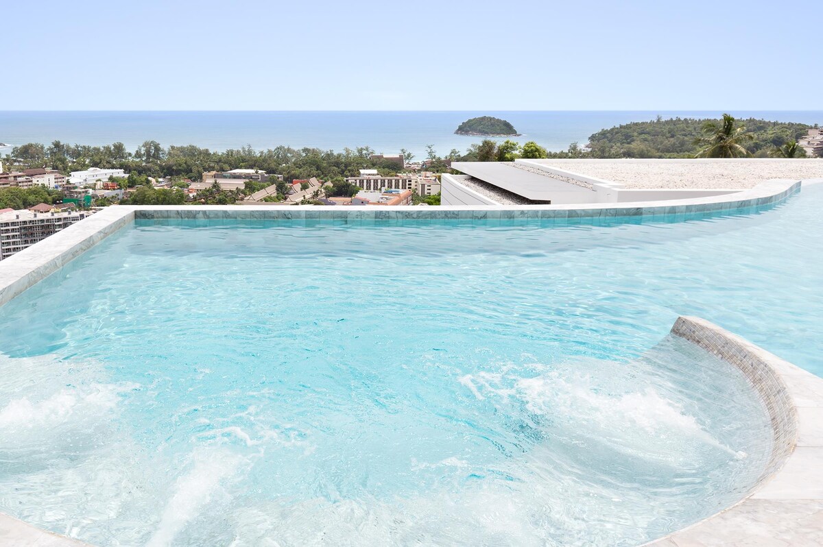 An infinity pool extends toward the horizon, offering sweeping views of the ocean and surrounding landscape. The clear water reflects the sky, while the edge of the pool gently curves, enhancing the sense of tranquility. Lush greenery and distant islands are visible in the background.