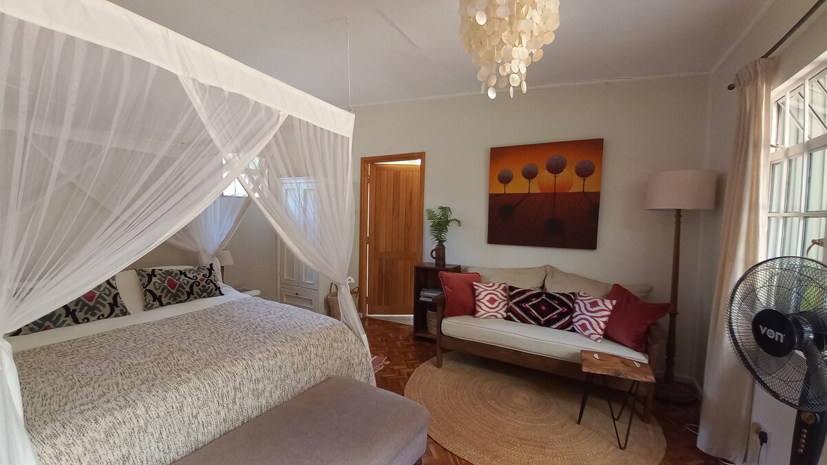 An open plan living space features a spacious bed draped with a canopy. A lounge area is equipped with a light-colored sofa adorned with decorative pillows and a round rug. Natural light filters through the window, enhancing the calming atmosphere.