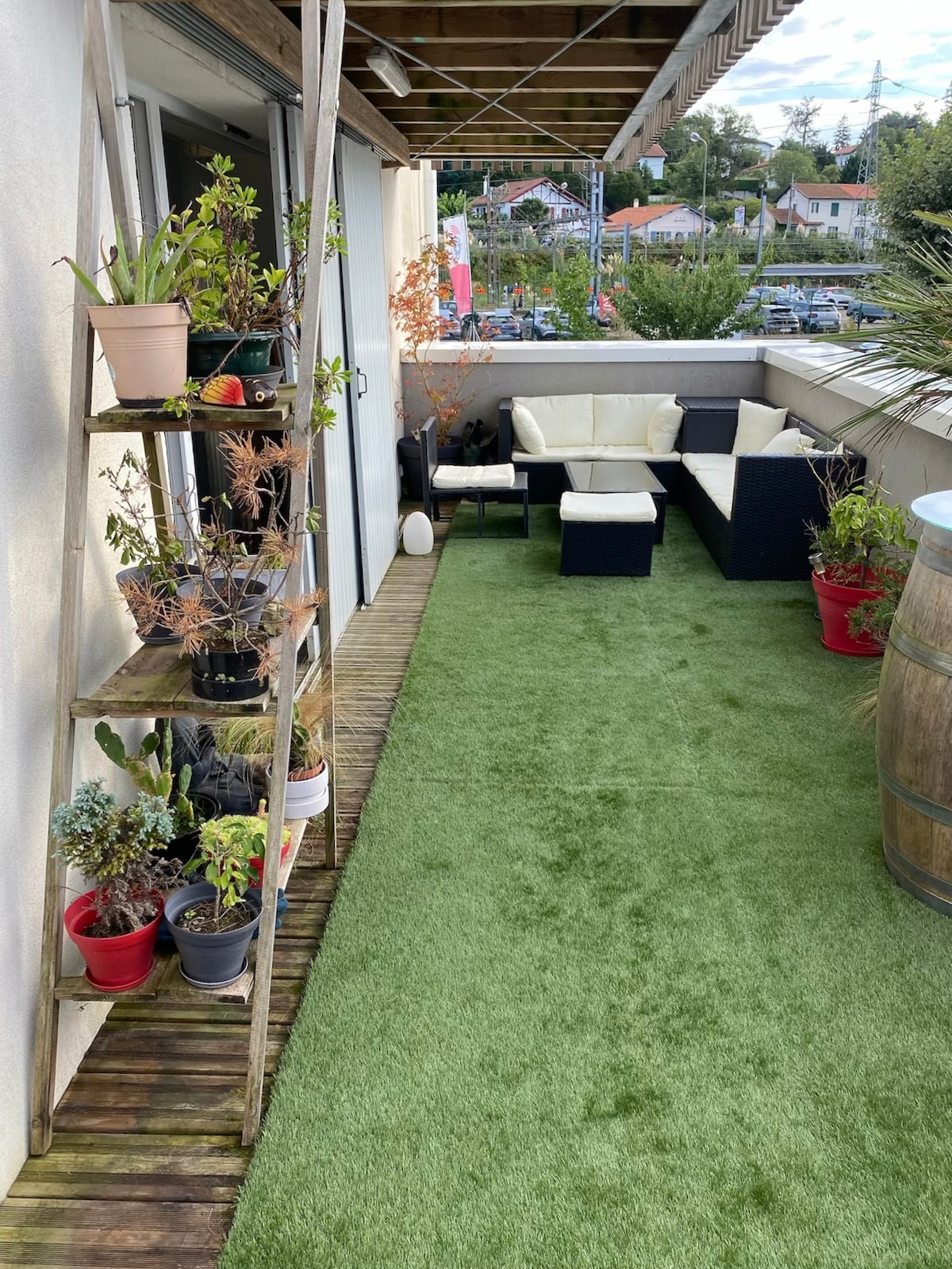 A private terrace is visible, featuring artificial grass and a selection of potted plants. Comfortable seating is arranged in a lounge layout, complemented by rustic barrel decor. The area provides a relaxed outdoor setting with views of the surroundings.