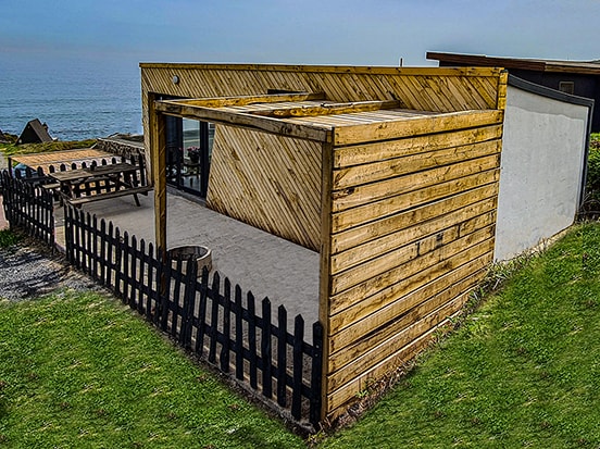 A wooden bungalow is set against a coastal backdrop, showcasing a spacious terrace with a light-colored surface. The exterior features natural wood paneling and a black picket fence, while lush greenery surrounds the structure, inviting relaxation with sea views.