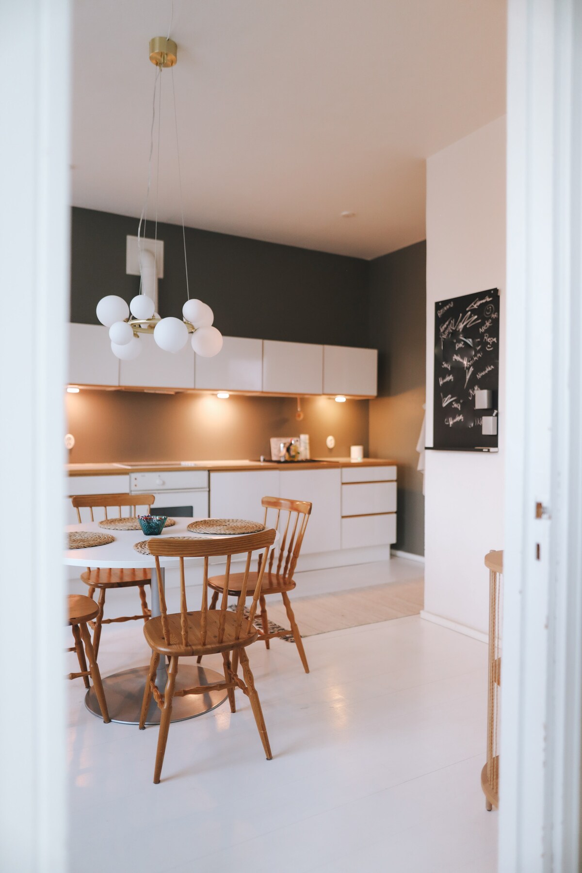 The kitchen features modern cabinetry and a circular dining table surrounded by wooden chairs. Warm lighting illuminates the space, highlighting the sleek design and neutral color palette. A decorative wall art piece adds character to the area.