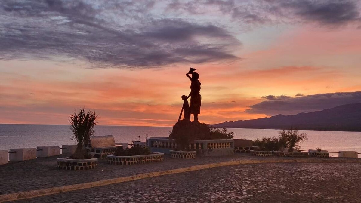 A statue of a figure is positioned against a backdrop of a colorful sunset. The silhouette stands prominently, with the ocean visible in the background. The ground is made of stone, and various plants can be observed around the area, contributing to the serene setting.