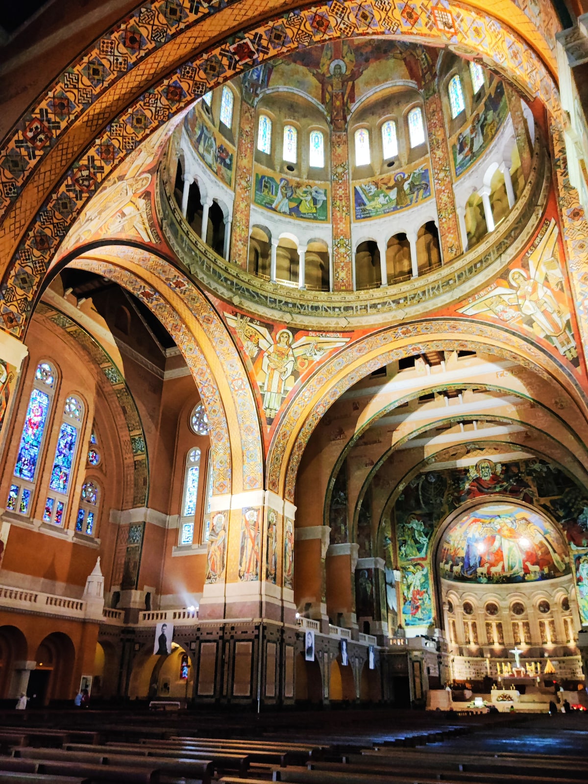 The interior of a grand cathedral is depicted, featuring soaring arches and intricate frescoes. Stained glass windows filter colorful light, illuminating the serene space. A large altar is visible at the far end, framed by detailed murals, enhancing the cathedral's spiritual ambiance.