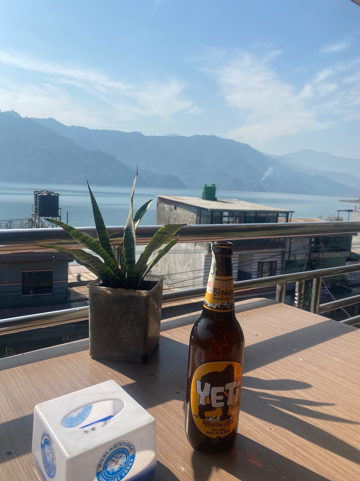 A table is set with a bottle of Yeti beer and a plant in a small pot. The backdrop features a serene lake surrounded by mountains, with clear skies above and gentle sunlight streaming down.