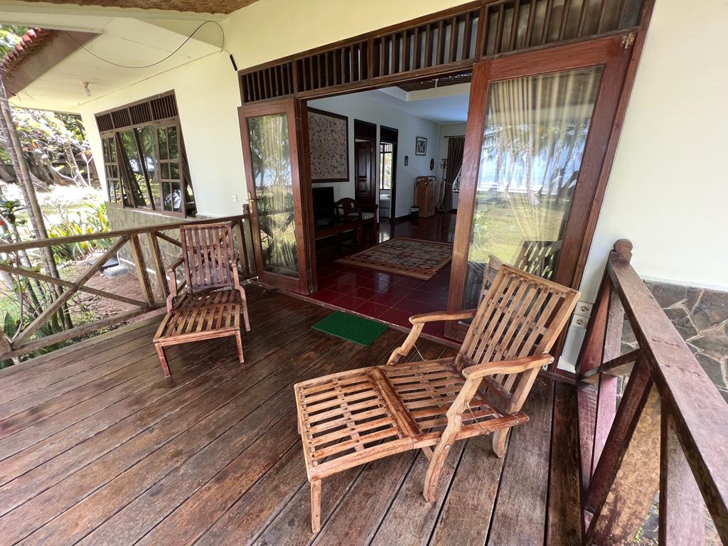 A welcoming porch area features two wooden chairs positioned on a wooden floor, with a view of the surrounding greenery. Sliding glass doors lead into the interior space, where natural light brightens the living area visible through the opening.