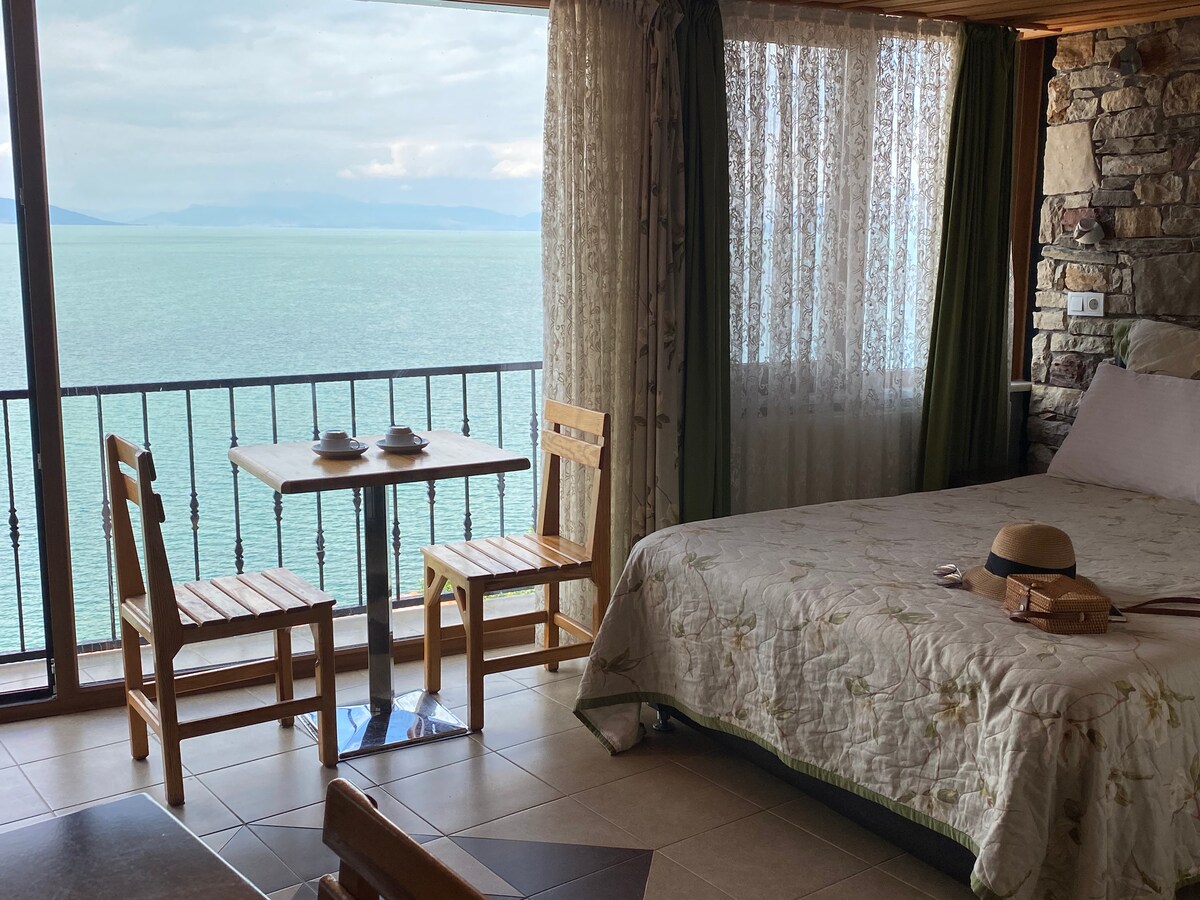 A cozy room is featured with a spacious bed and a small table set with two chairs, overlooking a stunning view of the lake. Light filtering through sheer curtains enhances the natural stone and wood finishes, creating a serene ambiance.