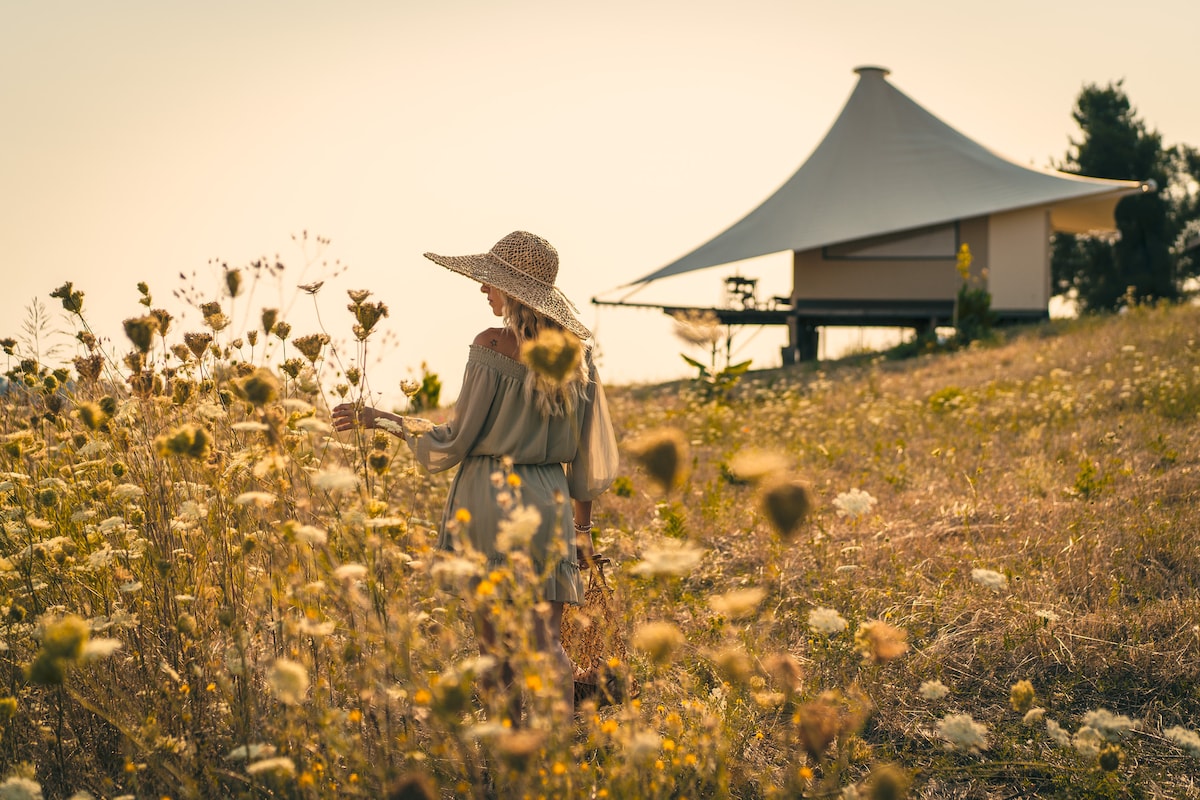 A woman in a wide-brimmed hat stands amidst tall wildflowers, gently touching the blossoms. In the background, a luxurious tent is positioned on a gentle hill, blending harmoniously with the natural landscape and warm evening light.