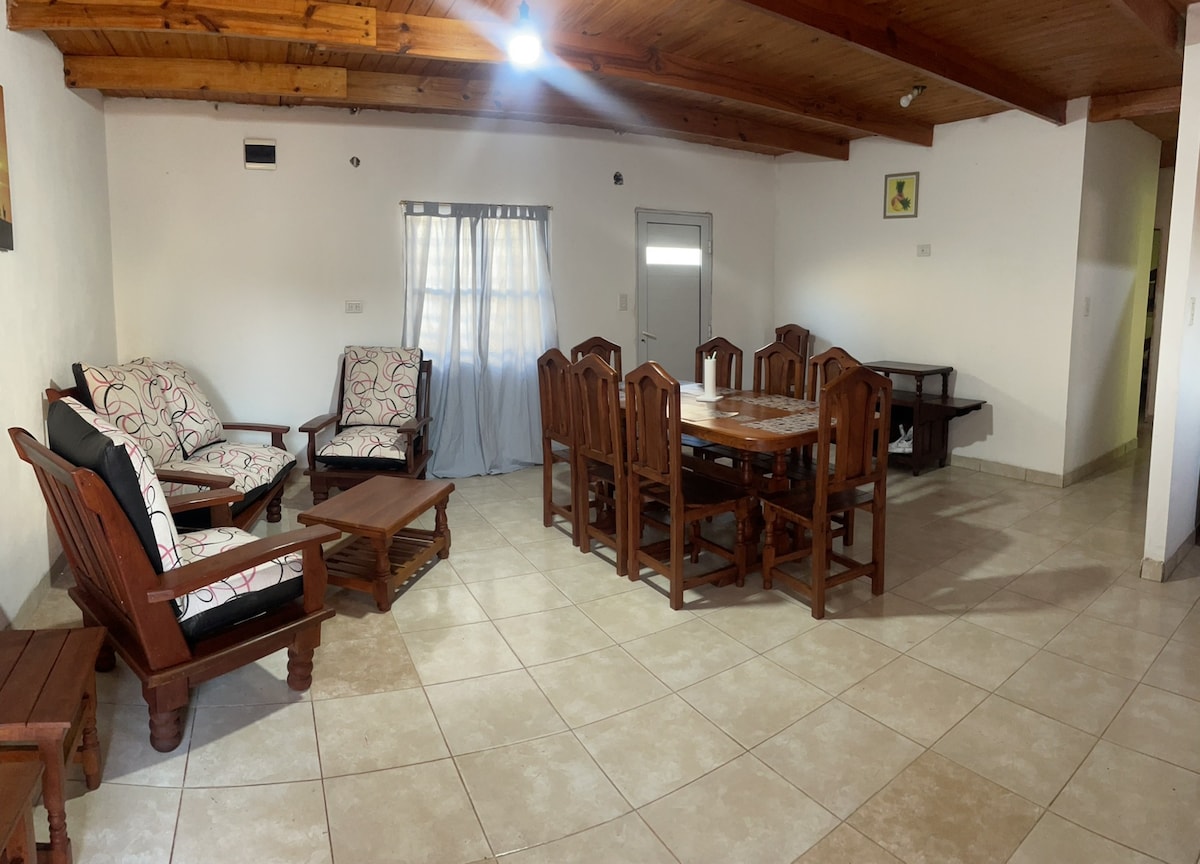 A spacious living and dining area is highlighted by wooden furniture, including a large dining table surrounded by chairs and a cozy seating arrangement. Natural light enters through a window with soft drapes, while the ceiling features wooden beams.