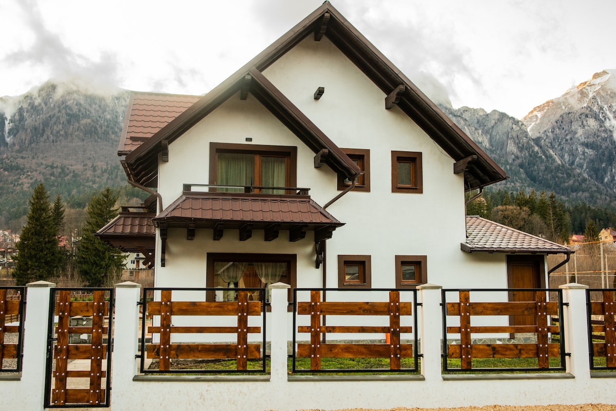 A modern two-story cottage is framed by wooden fencing, showcasing a combination of white walls and dark wood accents. The structure features a pitched roof and multiple windows, with mountains visible in the background under a partly cloudy sky.
