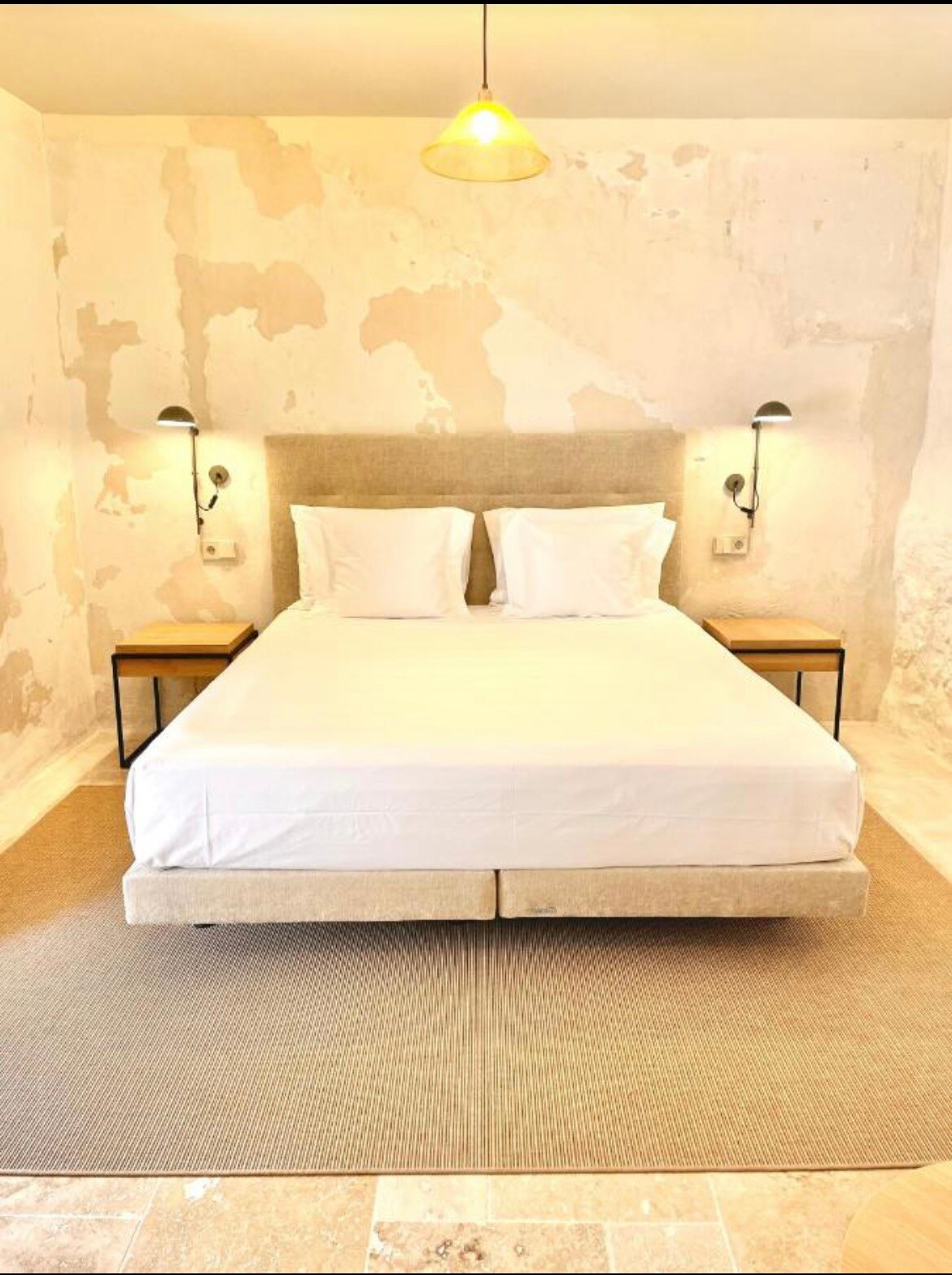 A spacious sleeping area features a comfortable bed with white linens, flanked by two bedside tables. Soft lighting is provided by a hanging bulb overhead. The walls display a textured finish, and a neutral-toned rug lies beneath the bed, creating a serene atmosphere.