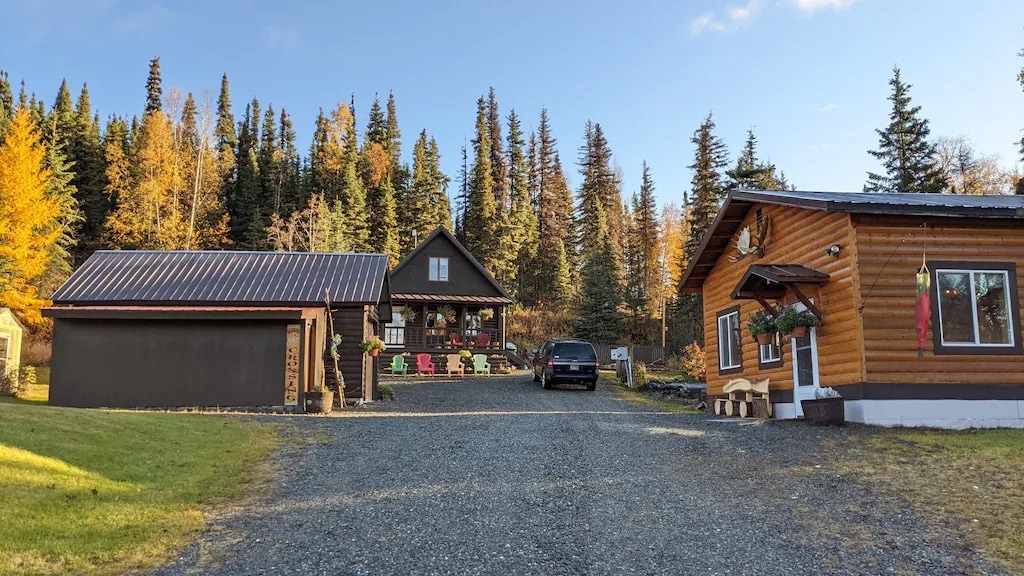 A gravel driveway leads to two rustic buildings surrounded by a natural landscape. A log cabin with a welcoming porch sits alongside a wooden guest house, both framed by vibrant autumn foliage. Parking space is available in front, with a few colorful chairs arranged outside.