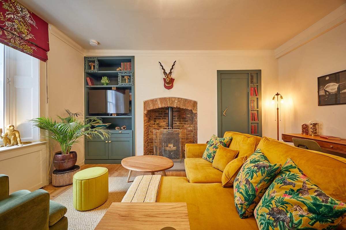 The inviting lounge area showcases a mustard-colored sofa adorned with patterned cushions and a round wooden coffee table. An exposed brick fireplace and a Freeview TV are visible, complemented by a shelf containing books and decorative items, enhancing the cozy ambiance.