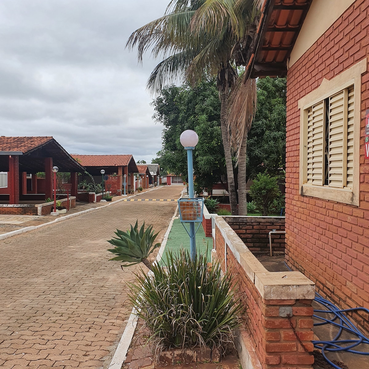A quiet pathway is seen leading through a well-maintained community, lined with brick and wooden chalets. Decorative palm trees and plants enhance the serene environment, while soft lighting from street lamps adds a touch of warmth to the scene.