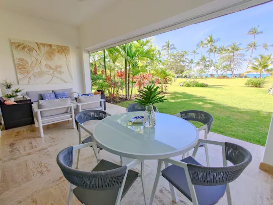 A bright patio area features a circular dining table surrounded by four chairs, set against a backdrop of lush greenery and palm trees. The floor-to-ceiling open window allows natural light to flood the space, while offering a view of the well-maintained garden and distant beach.