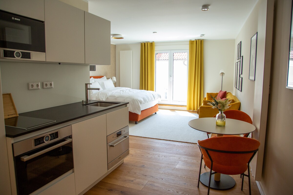 An open layout reveals a modern kitchen area with sleek appliances. A spacious bed is positioned beside large windows adorned with yellow curtains. A cozy seating area features an orange armchair and a small dining table that complements the welcoming design.