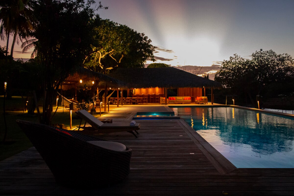A spacious deck surrounds a tranquil swimming pool, illuminated by soft lighting. A cozy lounge chair sits at the edge, while a thatched-roof bar area and seating are visible in the background. The lush landscape is complemented by a fading sunset sky.